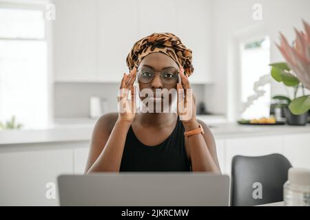 Afrikanische Frau, die eine Brille hält. Blick auf die Kamera, während Sie von zu Hause aus sitzen und arbeiten. Tragen Sie einen traditionellen Kopftuch Stockfoto