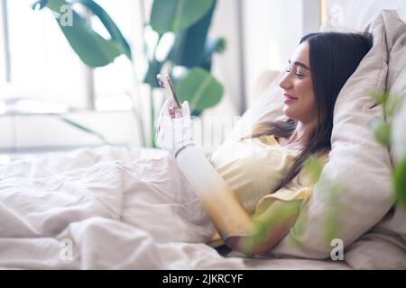 Mädchen mit bionischem Arm, das Telefon benutzt, soziale Medien liest und im Bett chattet, eine Frau mit Behinderung führt ein normales Leben mit künstlichen Prothesen Stockfoto