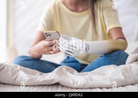 Mädchen mit bionischem Arm, das Telefon benutzt, soziale Medien liest und im Bett chattet, eine Frau mit Behinderung führt ein normales Leben mit künstlichen Prothesen Stockfoto
