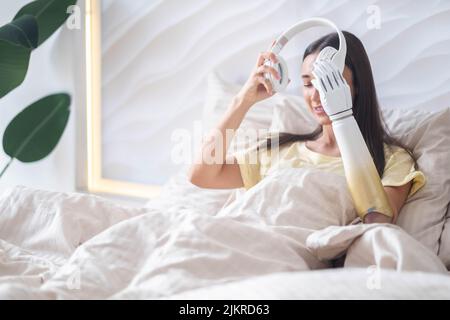 Glückliches Mädchen mit bionischen Arm entspannend Musik hören Kopfhörer im Bett zu Hause, Frau mit Behinderung führt normales Leben künstlichen Prothesen Extremität Stockfoto