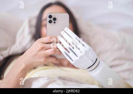 Mädchen mit bionischem Arm, das Telefon benutzt, soziale Medien liest und im Bett chattet, eine Frau mit Behinderung führt ein normales Leben mit künstlichen Prothesen Stockfoto