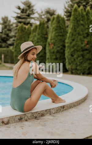 Nette junge Frau, die am Pool mit Handy im Hinterhof des Hauses sitzt Stockfoto