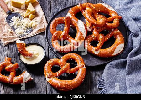 Weiche Brezeln in Form von Knoten gebacken und mit Salz auf schwarzem Teller auf dunklem Holztisch mit Käsesauce bestreut, horizontale Ansicht von oben Stockfoto