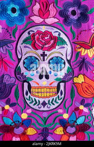 Handgeperlte mexikanische Damenhandtasche, hergestellt in Chiapas, mit einem Totenkopf und Blumen in verschiedenen Farben, mit violettem Hintergrund Stockfoto