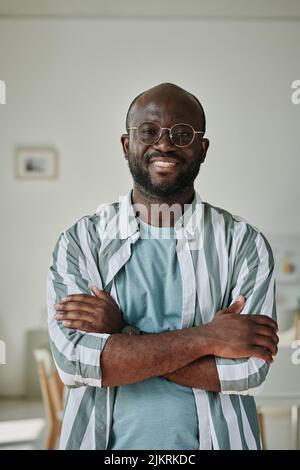 Porträt eines jungen afrikanischen Geschäftsmannes in einer Brille, der mit gekreuzten Armen steht und lächelt, während die Kamera im Büro steht Stockfoto
