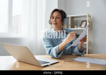 Eine aufmerksame, lächelnde, glückliche Schülerin in Kopfhörern, die von Online-Vorlesungen zu Hause lernt, schreibt die Zusammenfassung auf das Blatt. Glücklicher Büroangestellter ist da Stockfoto