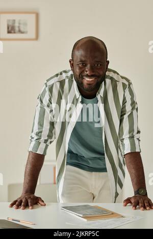 Porträt eines jungen afrikanischen Geschäftsmannes, der lächelt und die Kamera auf seinem Schreibtisch anlehnt Stockfoto