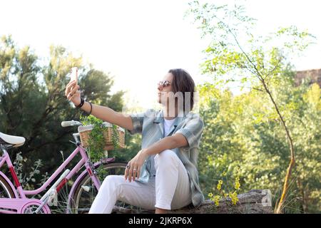Junger, gutaussehender Mann, der im Frühling ein Selfie im Park machte und sich mit seinem Fahrrad an der Seite erholte Stockfoto