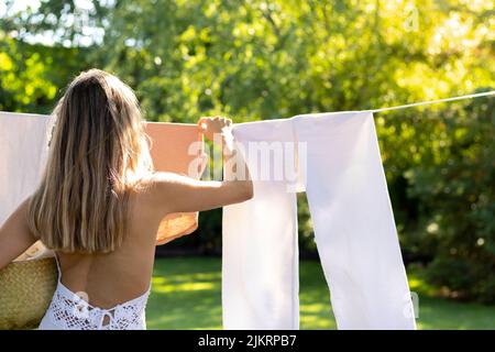 Rückansicht einer nicht erkennbaren jungen blonden Frau, die die Kleidung in einer Linie draußen im Garten aufhängte, umgeben von der Natur Stockfoto