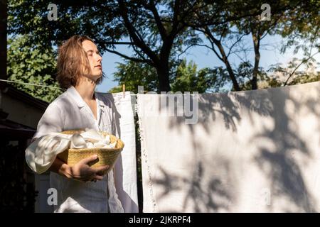 Junger Mann mit langen Haaren und geschlossenen Augen, der die Sommersonne auf seinem Gesicht aufhängt, während er die Wäsche aufhängt Stockfoto