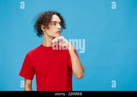 Nachdenklich nachdenklich unglücklich junge lockig gebräunt kaukasischen Kerl in grundlegenden roten T-Shirt posiert isoliert auf blauem Hintergrund. Lifestyle und Emotionen Stockfoto