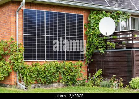 Sonnenkollektoren, die an ein Haushaus angeschlossen sind, um Platz zu sparen und grüne Energie zu produzieren. Schöner Garten mit Weinreben und Blumen wachsen. Stockfoto