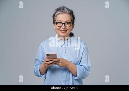 SMS oder Tippen Halten des Smartphones in den Händen reife grauhaarige Frau, die online arbeitet oder einkauft und in sozialen Medien nachcheckt. Hübsche Frau in blauer Bluse isoliert auf weißem Hintergrund. Stockfoto