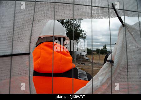 CHERTSEY, Surrey, Großbritannien. 3.. August 2022. Ein Sicherheitsbeamter am Eingang des Tunnels versucht, die Sicht eines Pressefotografen zu blockieren. Ein erfahrener Klimaaktivist, bekannt als Digger, gräbt derzeit einen Tunnel in Chertsey an Land, wo ExxonMobil eine neue Flugkraftstoff-Pipeline nach London Heathrow baut. Die Stop the Pipeline to Exxtinction-Gruppe fordert keine neuen Investitionen oder Erweiterungen in die Infrastruktur fossiler Brennstoffe. Quelle: Maureen McLean/Alamy Live News Stockfoto