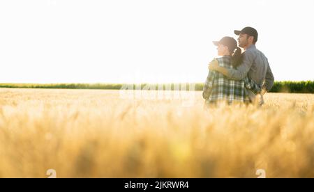 Ein paar Bauern in karierten Hemden und Kappen stehen bei Sonnenuntergang auf dem landwirtschaftlichen Weizenfeld umarmt Stockfoto