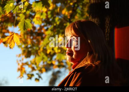 Unschärfe-Porträt der schönen kaukasischen blonde Frau in bunten gelben Ahorn Herbst Park. Helle stilvolle Frau im Mantel in der Nähe Baumstamm. Hallo Fall CO Stockfoto