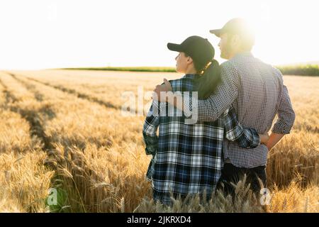 Ein paar Bauern in karierten Hemden und Kappen stehen bei Sonnenuntergang auf dem landwirtschaftlichen Weizenfeld umarmt Stockfoto