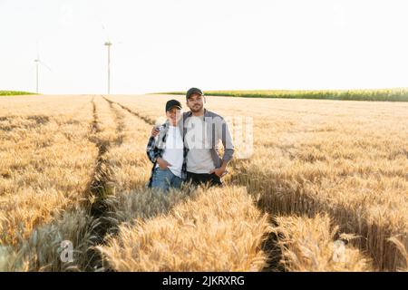 Ein paar Bauern in karierten Hemden und Kappen stehen bei Sonnenuntergang auf dem landwirtschaftlichen Weizenfeld umarmt Stockfoto