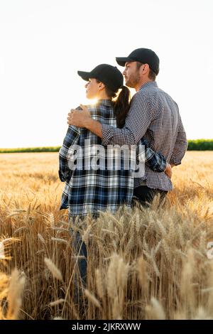 Ein paar Bauern in karierten Hemden und Kappen stehen bei Sonnenuntergang auf dem landwirtschaftlichen Weizenfeld umarmt Stockfoto