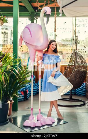 Eine junge schöne Frau mit langen dunklen Haaren in einem kurzen Oberteil steht neben einer großen Flamingo-Figur und spielt, posiert, lacht. Stockfoto