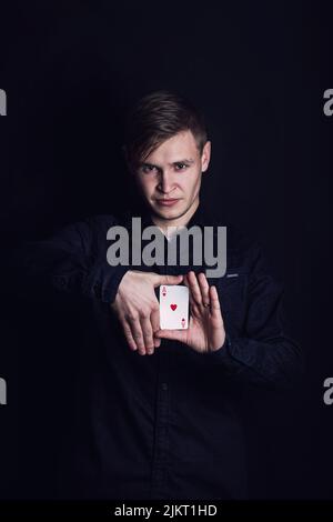 Selbstbewusster junger Mann, der Tricks mit Spielkarten zeigt, wie ein Zauberer, isoliert auf dunklem Hintergrund. Gewinner der Glücksspiele, erfolgreicher Spieler Stockfoto