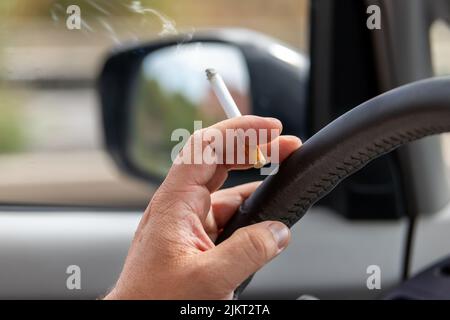 Nahaufnahme eines männlichen Fahrers, der tagsüber auf der Hauptstraße in der Stadt mit der Zigarette unterwegs war. Mann, der drinnen raucht Stockfoto