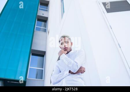 Von unten zeigt ein afroamerikanischer Mann in elegantem weißen Hemd, der die Kamera anschaut, während er am Sommertag in der Stadt gegen moderne Wolkenkratzer steht Stockfoto