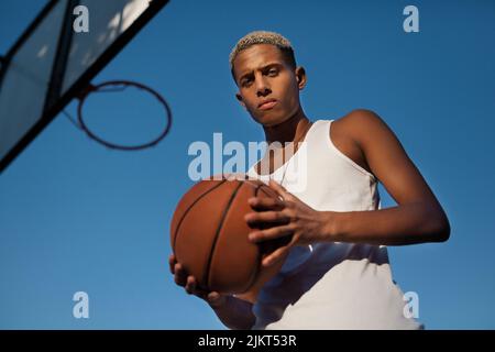 Von unten ein ernsthafter afroamerikanischer Basketballspieler mit Ball, der die Kamera in der Nähe des Reifens gegen den blauen Himmel auf dem Spielplatz anschaut Stockfoto