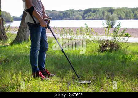 Junger Mann mit einem drahtlosen Metalldetektor auf der Suche nach einem Schatz am Flussufer. Bäume im Hintergrund Stockfoto