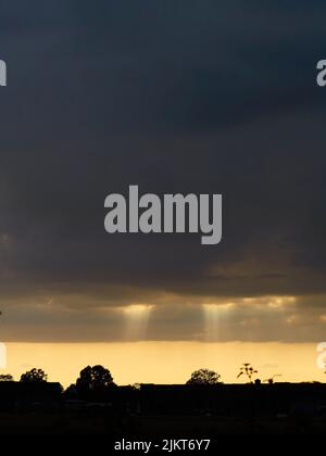 Skyscape mit Sonnenlicht, das durch die bleierne Düsternis von schweren Sturmwolken über silhouettierten Gebäuden und Bäumen platzt. Stockfoto