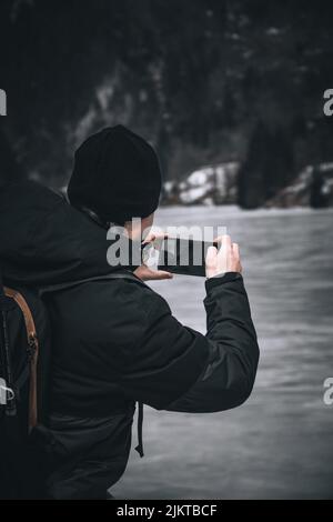 Vertikale Aufnahme einer Person von hinten, die schwarze Jacke und Mütze trägt und mit seinem Handy fotografiert Stockfoto