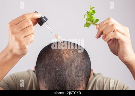 Junger Mann zeigt Minzblätter und tragen Haar ätherisches Öl in kahlen Flecken vor grauem Hintergrund. Stockfoto
