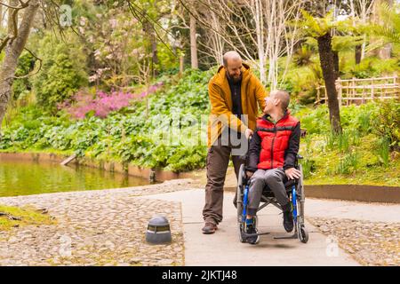 Gelähmter junger Mann in einem Rollstuhl, der von einem Freund in einen öffentlichen Stadtpark geschoben wird Stockfoto