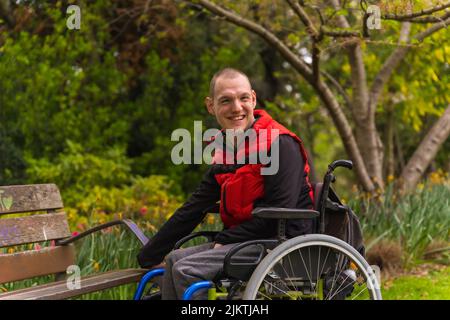 Porträt eines gelähmten jungen Mannes in einer roten Weste in einem öffentlichen Park in der Stadt. Im Rollstuhl neben einem Parkstuhl sitzen Stockfoto