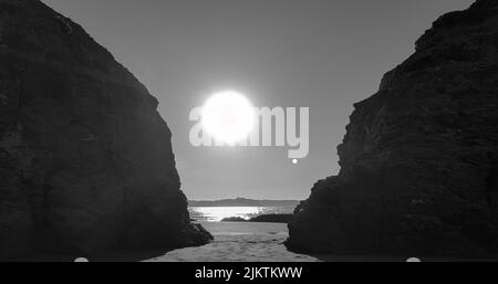 Eine Graustufe von Meer oder Ozeanküste mit Sonne, die ringsum leuchtet und massiven Felsen im Vordergrund Stockfoto