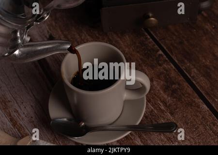 Metallvase gießt Kaffee in eine Keramikschale auf einem Holztisch Stockfoto