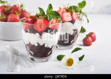 Kleinigkeiten. Brownies, frische Erdbeere und Mascarpone-Creme in Schichten auf Glas auf weißem Hintergrund gezackt. Portionsdesserts. Kein gebackener Brownie Cheesecak Stockfoto