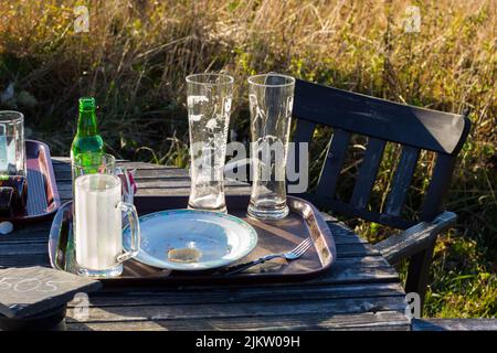 Leere Gläser und Teller in einem Metalltablett auf einem Holztisch bei einem Stuhl im Garten an einem schönen sonnigen Tag. Stockfoto