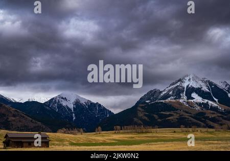 DER SCHNEE BEDECKTE DIE ABSAROKA-BERGKETTE IN PRAY MONTANA MIT EINEM SCHÖNEN, ÜPPIGEN FELD IM VORDERGRUND UND EINEM DUNKLEN, STÜRMISCHEN HIMMEL Stockfoto