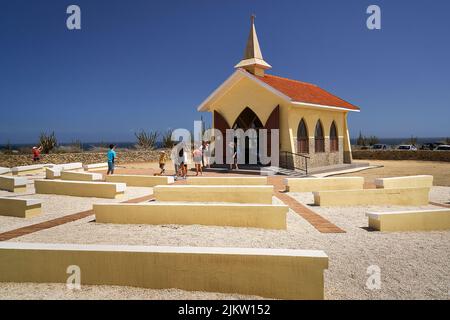 Besucher der leuchtend gelben Alto Vista Chapel und ihrer Außenbänke vor dem blauen karibischen Himmel. Stockfoto