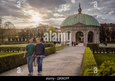 Zwei junge Freunde gehen durch den Hofgarten, den Park in der Münchner Innenstadt. Episch atemberaubend schöner Sonnenuntergang im Hintergrund des Stadtparks Stockfoto