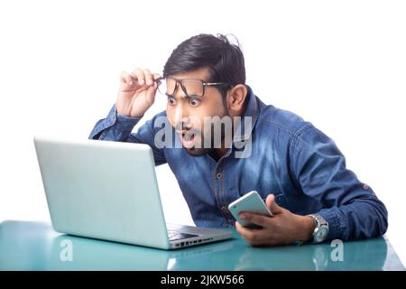 Erstaunter Geschäftsmann, der auf den Laptop-Bildschirm schaute, indem er die Brille hob und das Mobiltelefon auf weißem Hintergrund an der Hand hielt Stockfoto