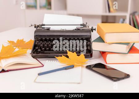 Es gibt Retro-Schreibmaschine auf weißem Tisch. In der Nähe befinden sich Bücher, Notizbuch mit Stift, Mobiltelefon und gelbe Ahornblätter. Arbeitsplatz des Schriftstellers. Stockfoto