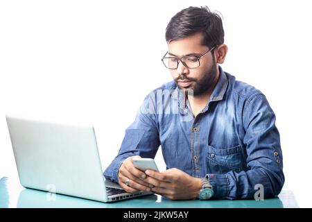 Abgelenkter junger Mann, der vor einem weißen Laptop auf dem Handy tippt Stockfoto