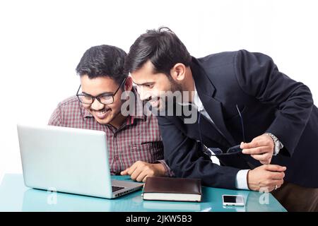 Glücklicher indischer Geschäftsmann im Gespräch mit einem Kollegen, der mit einem Laptop auf weißem Hintergrund zusammensitzt Stockfoto
