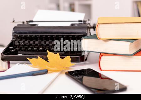 Es gibt Retro-Schreibmaschine auf weißem Tisch. In der Nähe befinden sich Bücher, Notizbuch mit Stift, Mobiltelefon und gelbe Ahornblätter. Arbeitsplatz des Schriftstellers. Stockfoto