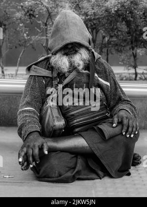 Eine vertikale Graustufenaufnahme eines obdachlosen tamilischen indischen Mannes, der auf dem Bürgersteig in Kuala Lumpur, Malaysia, sitzt Stockfoto