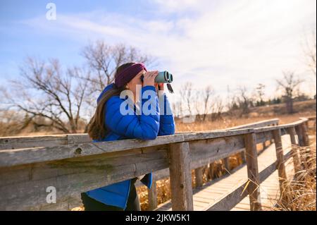 Eine Frau in einer blauen Daunenjacke blickt an einem sonnigen Frühlingstag durch ein blaues Fernglas auf einer Brücke in einem Park in die Ferne. Stockfoto