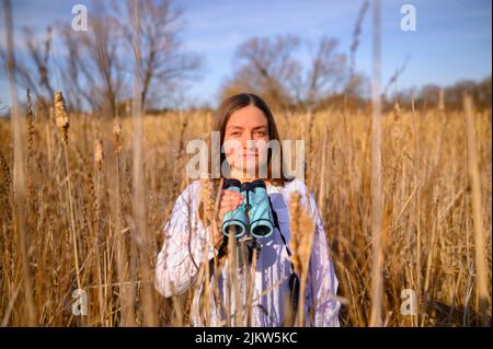 Frau, die die Kamera direkt anschaut, mit neutralem Ausdruck, mit einem blauen Fernglas, das tagsüber in einem Feld von Katzenschwänzen in einem Sumpfgebiet steht. Stockfoto