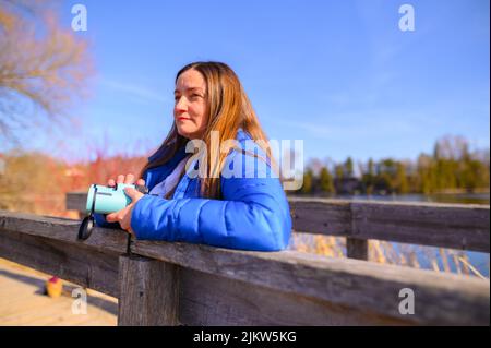 Eine Frau in einer blauen Daunenjacke blickt an einem sonnigen Frühlingstag auf einer Brücke mit einem Fernglas in einem Park in die Ferne. Stockfoto